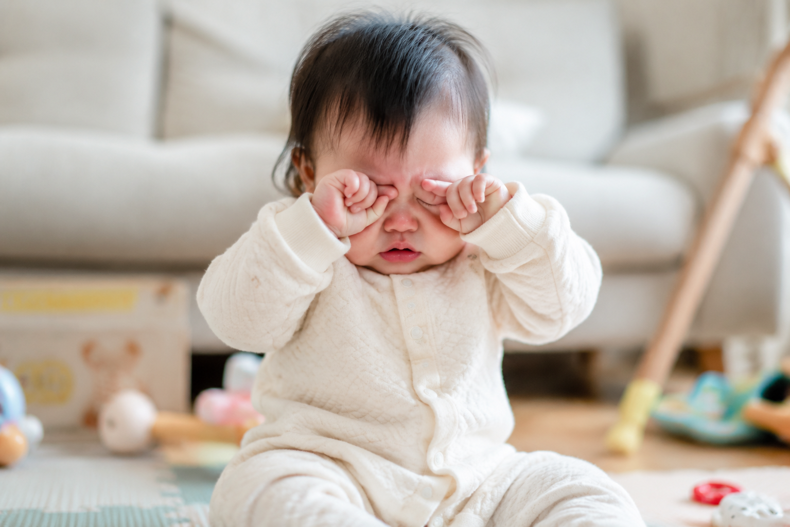 A cute 8-month-old Chinese baby sitting on a soft play mat in a cozy living room, rubbing their eyes with tiny hands, slightly red nose, looking a bit uncomfortable but extremely cute. Bright indoor natural lighting, soft pastel colors, photorealistic, 8k resolution, shot with an 85mm portrait lens, highly detailed face. --ar 3:4