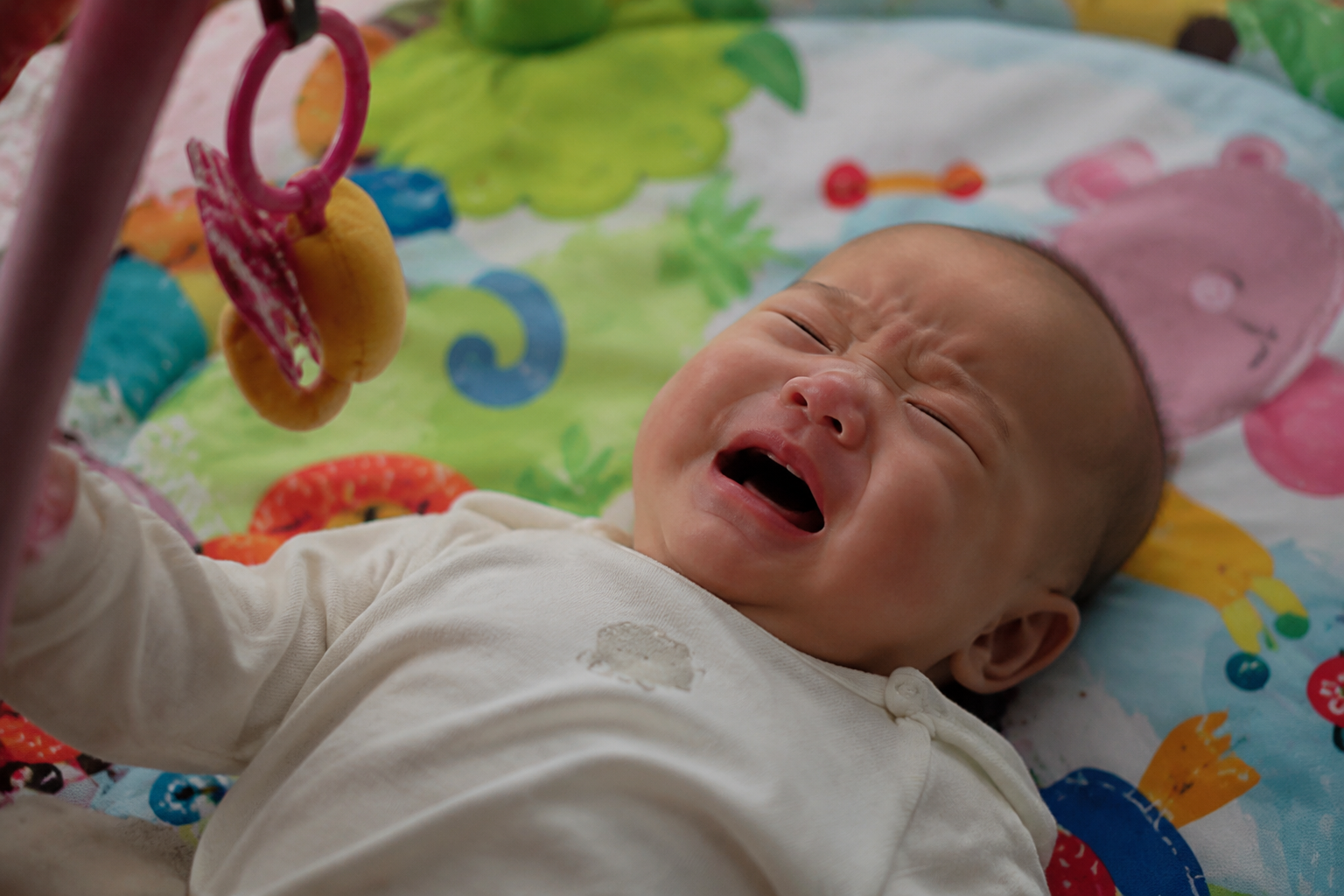 A cute 3-month-old Chinese baby lying on a colorful baby play mat, looking fussy and crying gently, humid and gloomy indoor lighting typical of rainy season, realistic photography, soft lighting, 8k resolution, photorealistic, cinematic light, highly detailed.