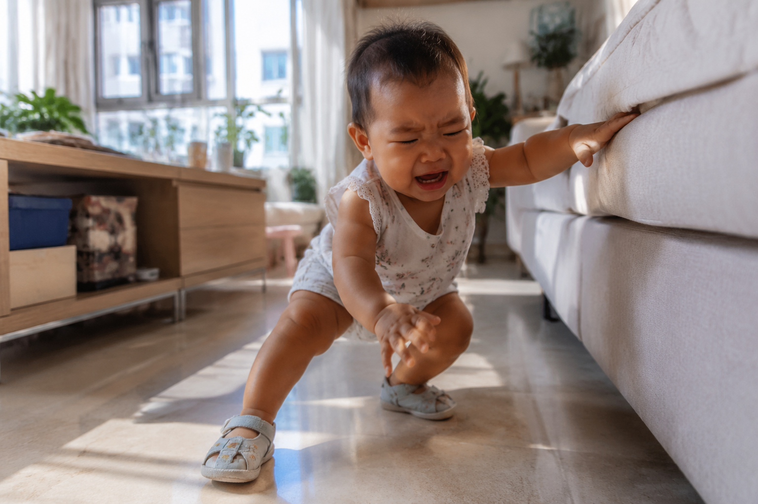 A cute 11-month-old Chinese baby learning to walk in a cozy living room, wearing cute summer clothes and ill-fitting soft sole baby sandals or crocs, the baby is slightly losing balance and about to fall while holding onto a sofa, frustrated expression, soft natural sunlight from the window, cinematic lighting, ultra-realistic, 8k resolution, photorealistic, photography by Sony A7R IV.