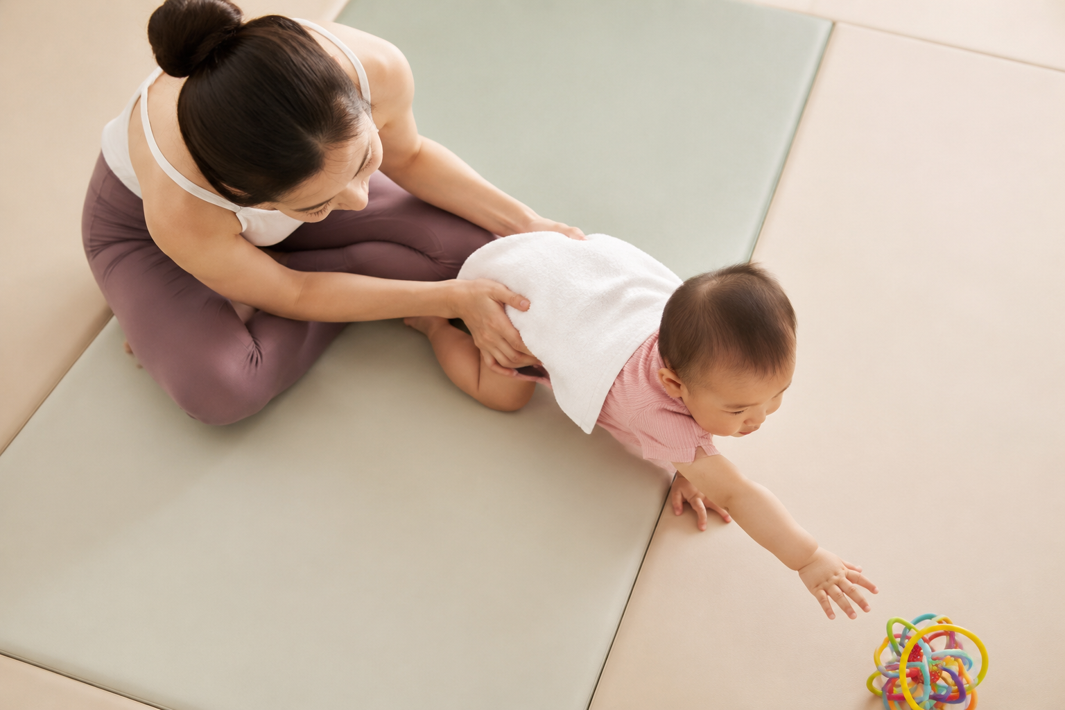 Top-down angle photography of a young mother wearing comfortable yoga clothes, gently supporting her baby's belly with a soft white bath towel to help the baby practice crawling on a padded floor mat. The baby is reaching for a colorful toy. Bright indoor lighting, warm pastel colors, hyper-realistic, parenting magazine style. --ar 3:4 --v 6.0