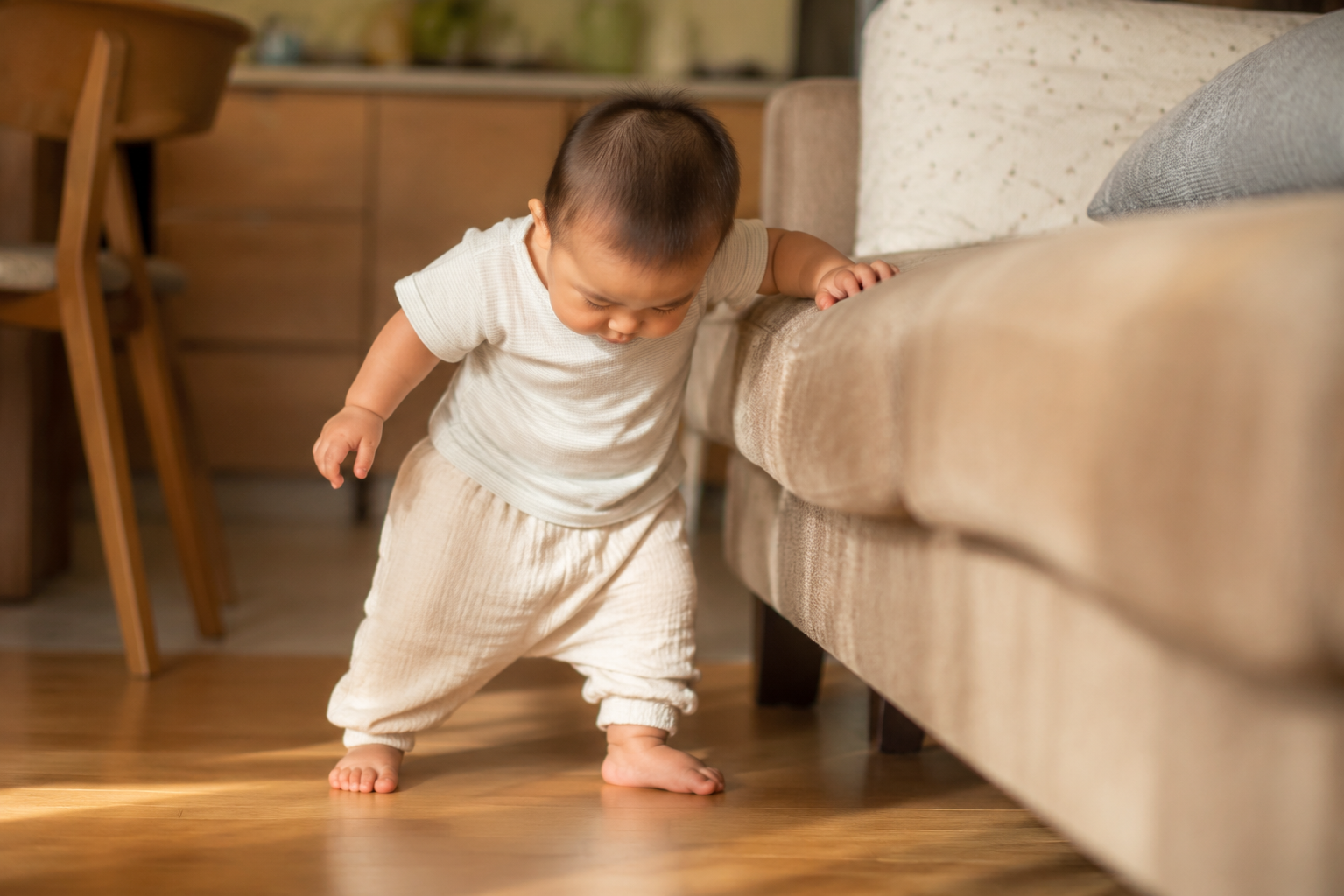 A cute 10-month-old Chinese baby boy trying to stand up holding a living room sofa, looking down at his feet. Warm indoor sunlight, soft cinematic lighting, ultra-realistic, 8k resolution, photorealistic, adorable and heartwarming atmosphere --ar 3:4