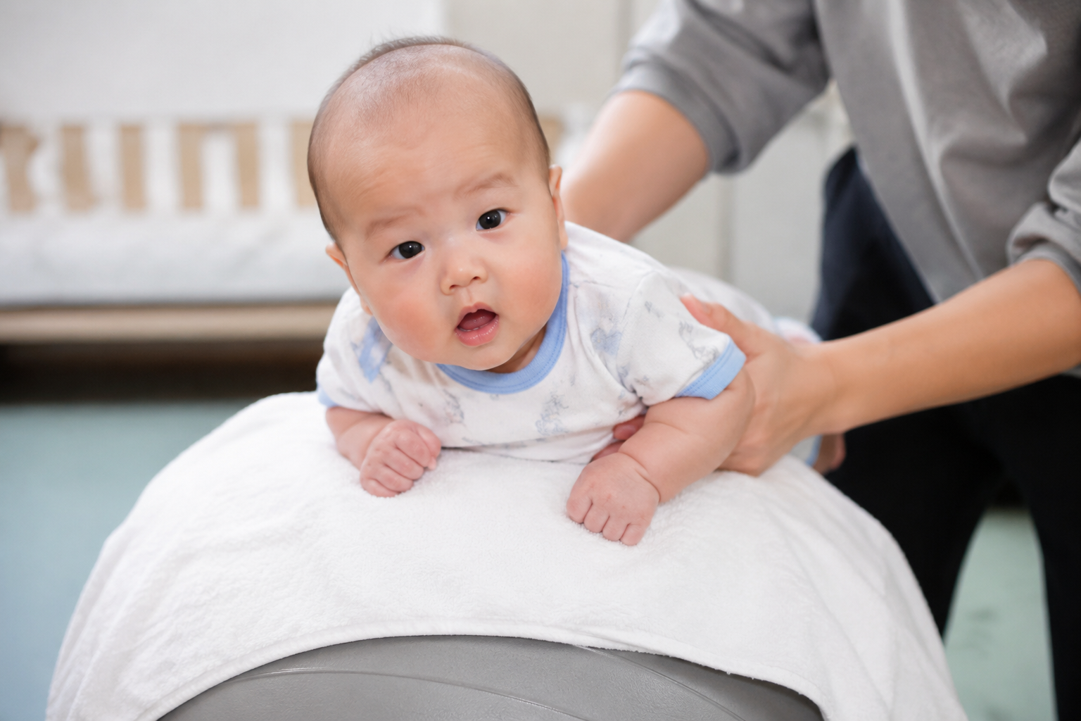 A 3-month-old Chinese baby lying on stomach on a large fitness yoga ball covered with a soft white towel, Chinese parent's hands gently and securely holding the baby's sides, safe and engaging environment, bright indoor lighting, realistic photography, 8k, sharp focus, magazine photography.