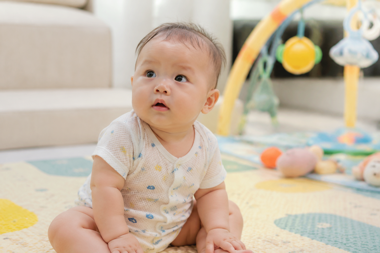 A cute 6-month-old Asian baby sitting on a soft play mat, wearing a light summer onesie, slightly sweating on the forehead, looking up curiously. Soft bright indoor lighting, cozy home environment, pastel colors, realistic photography, 8k resolution, photorealistic, shot on 35mm lens --ar 3:4