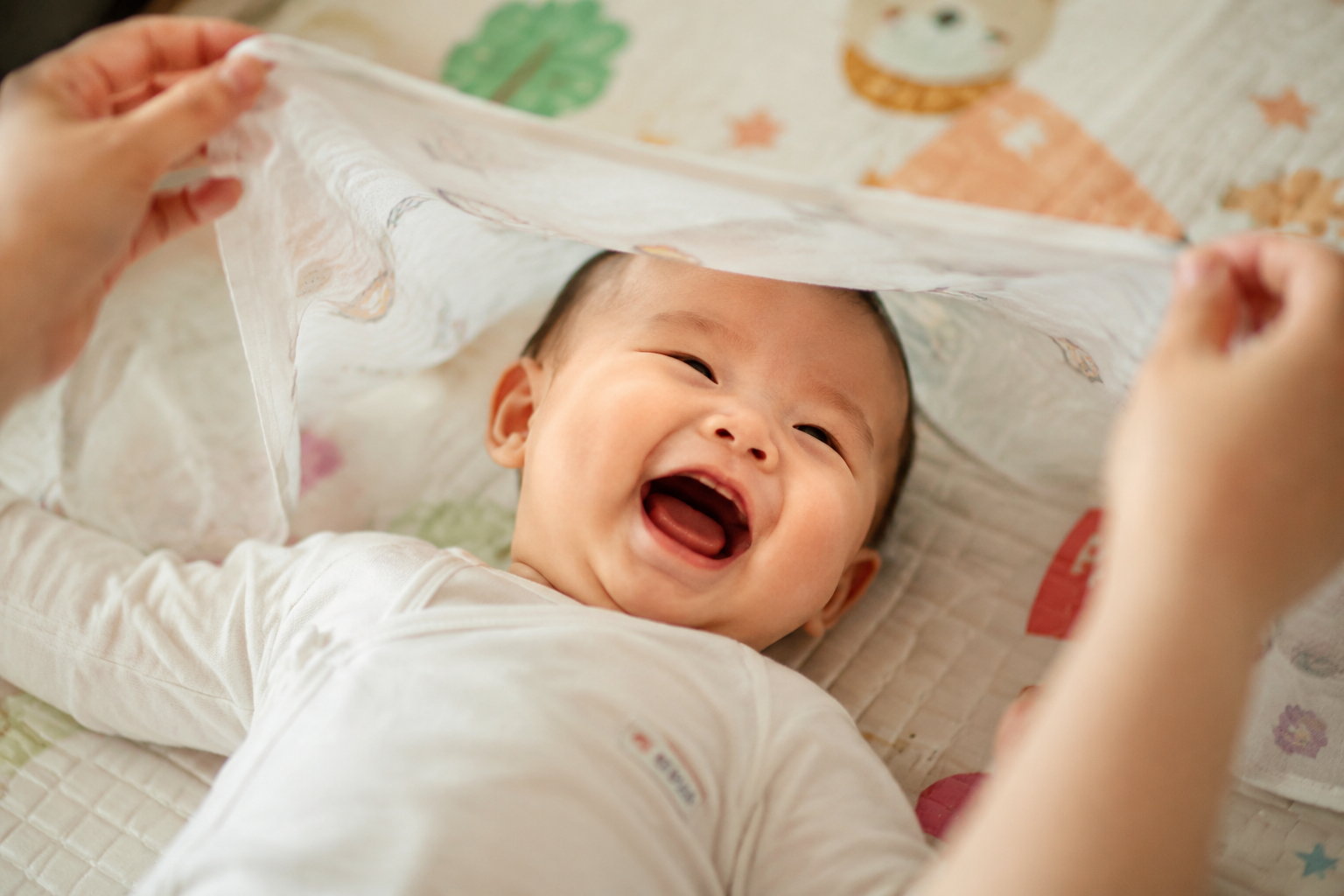 A heartwarming scene of a 5-month-old Chinese baby laughing joyfully as a sheer, light cotton gauze blanket is gently lifted off their face. The baby is lying on a soft play mat. The mother's hands are visible holding the edges of the blanket. Soft natural light, high detail, capturing pure joy, emotional photography, 85mm lens.