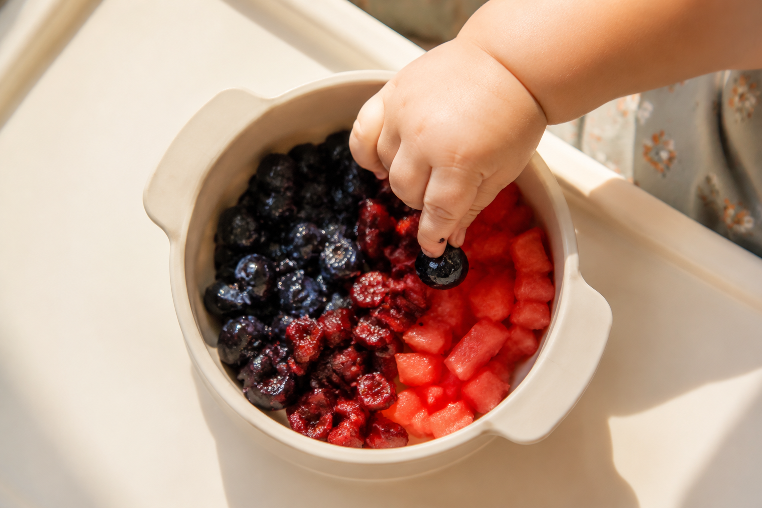 Top-down view, a silicone suction bowl on a baby high chair tray. Inside the bowl, there are squished blueberries, finely diced cherries without pits, and small watermelon sticks. A chubby little hand of a Chinese baby is using the thumb and index finger (pincer grasp) to pick up a squished blueberry. Bright morning sunlight, macro photography, high quality food photography, vibrant colors, shallow depth of field. --ar 1:1