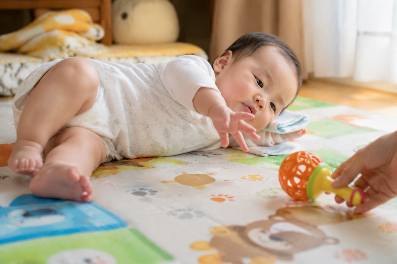 Realistic photography, A chubby 5-month-old Chinese baby lying on their back on a bright play mat, trying to reach a bright rattling toy placed on their right side on the floor. The baby is twisting their hip and lifting their shoulder, halfway into a rolling motion. A gentle Chinese mother's hand is visible holding the toy. Soft natural window light, warm and inviting atmosphere, ultra-detailed, 8k, photorealistic.