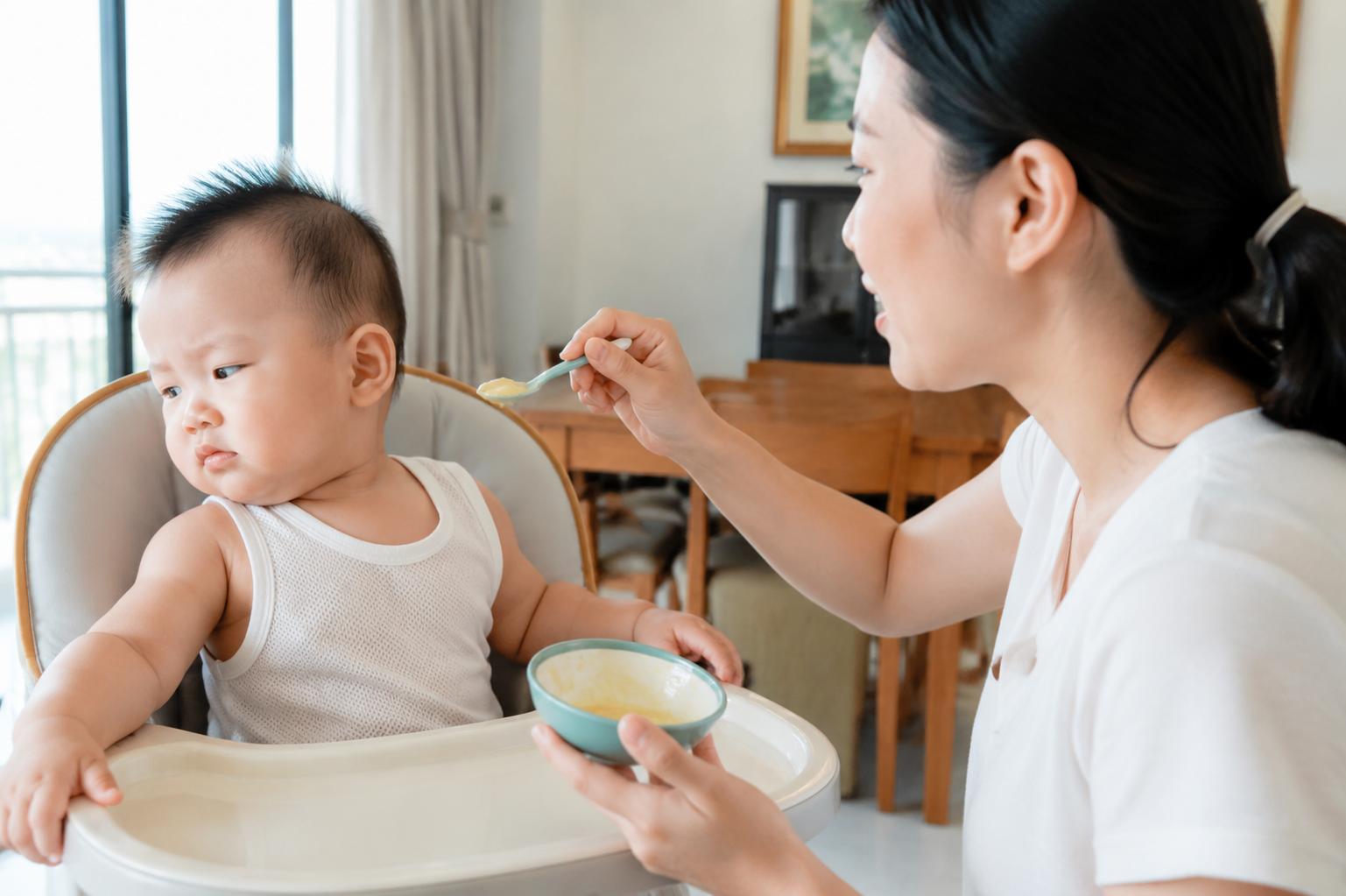 A cute 6-month-old Chinese baby sitting in a baby high chair, turning head away and refusing to eat baby food from a spoon held by a mother. Summer clothing, bright natural lighting, indoor dining room, highly detailed, photorealistic, Midjourney style, 8k resolution.