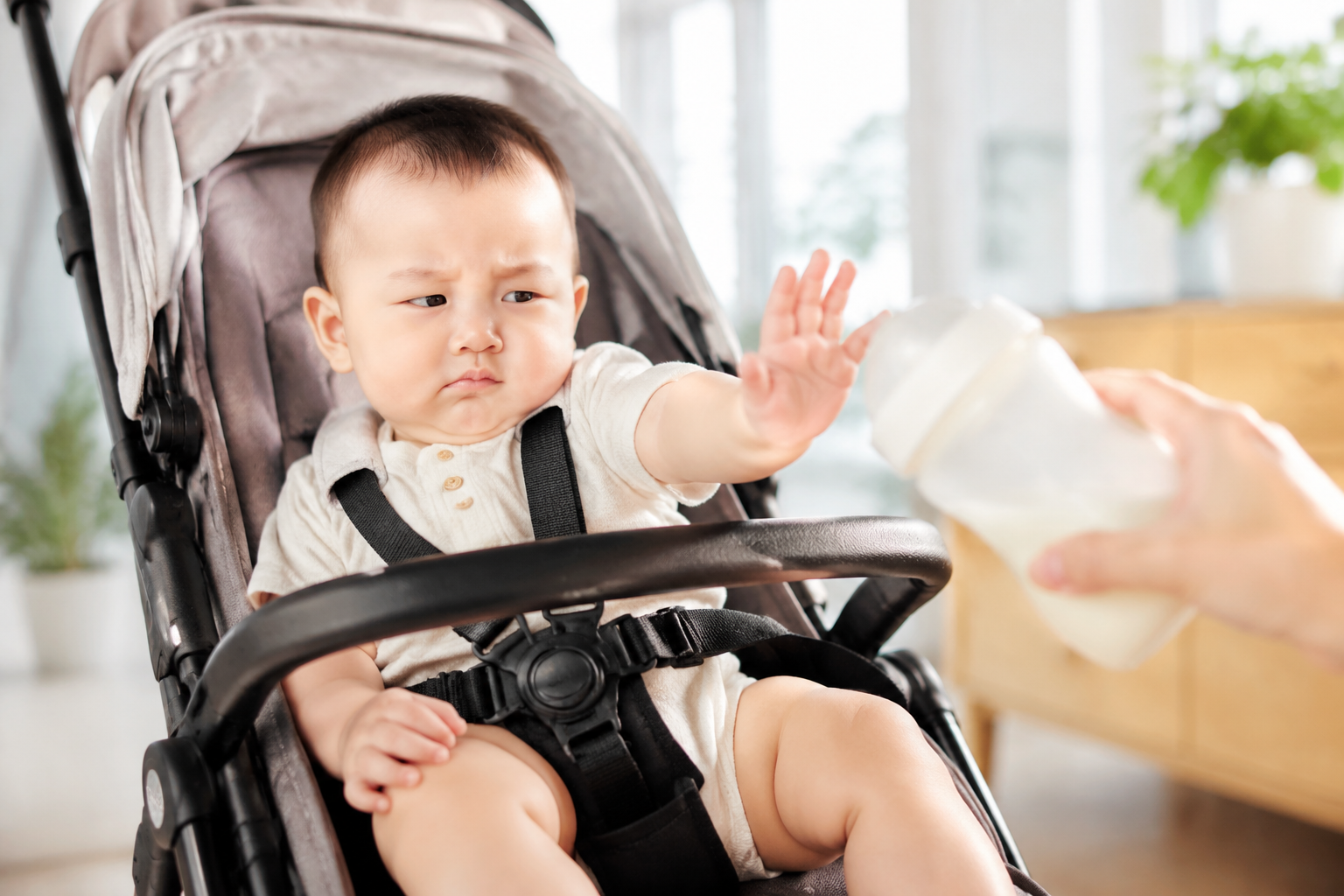 A cute 8-month-old Chinese baby sitting in a baby stroller indoors on a bright summer day, pushing away a milk bottle with a slightly fussy and funny expression. Soft natural lighting from the window, warm summer tones, high-resolution photography, photorealistic, 8k, soft focus background.
