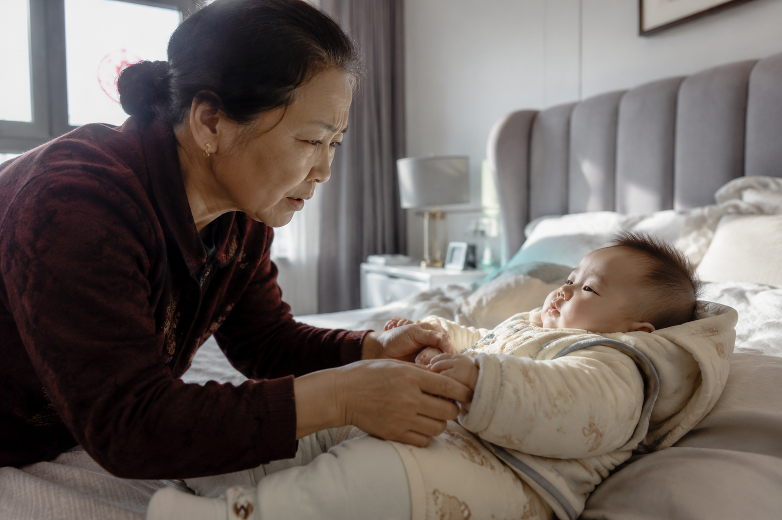 A photorealistic image of a concerned Chinese grandmother touching the hands of a cute 6-month-old Chinese baby. The baby is lying on a bed, wearing too many layers of thick clothes in a bright, sunlit modern bedroom. The grandmother looks worried. Soft natural lighting, highly detailed, 8k resolution, shot on 35mm lens, cinematic lighting.
