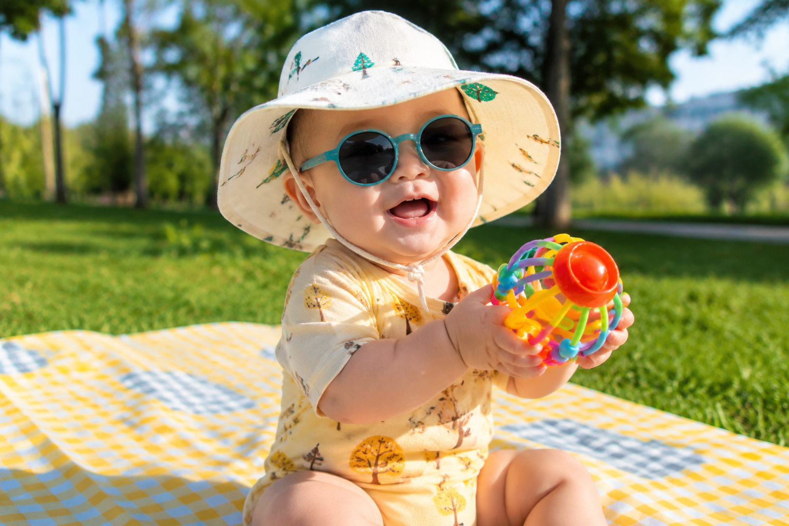 A hyper-realistic, bright and cheerful photo of a happy 10-month-old Chinese baby sitting on a picnic mat on green grass. The baby is wearing a cute wide-brimmed sun hat (UPF 50+) and stylish, colorful silicone baby sunglasses. The baby is smiling brightly, actively playing with a toy. Warm morning sunlight, shallow depth of field, vibrant colors, 8k, ultra-detailed, photorealistic. --ar 3:4