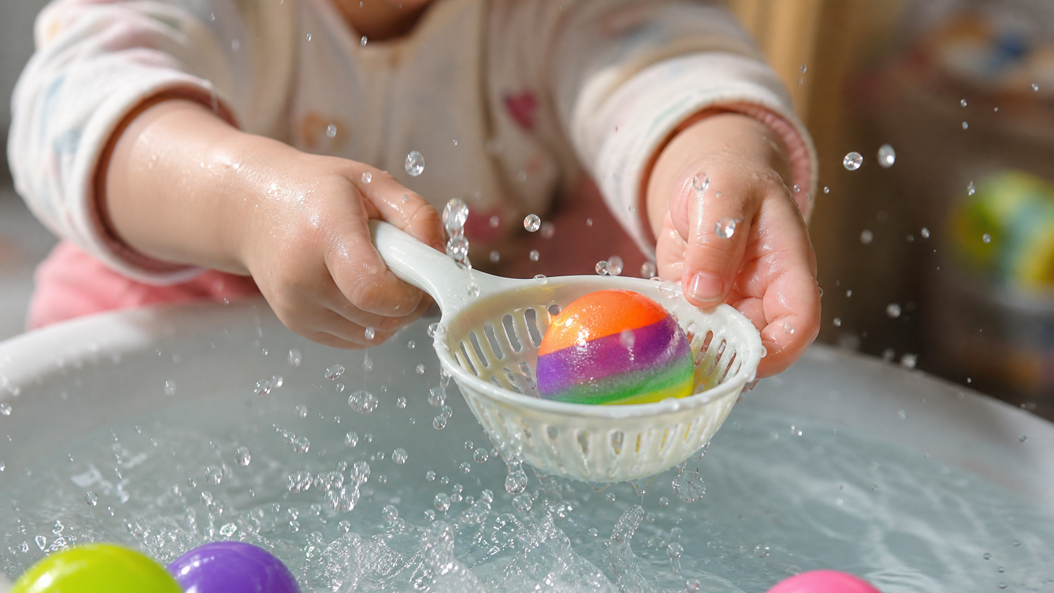 Close up of a cute Chinese toddler's little hands holding a small plastic strainer, trying to catch a colorful floating ball in a basin of water. Splashing water droplets, high-speed photography, bright indoor lighting, highly detailed, photorealistic, depth of field, warm and cheerful vibe --ar 3:4