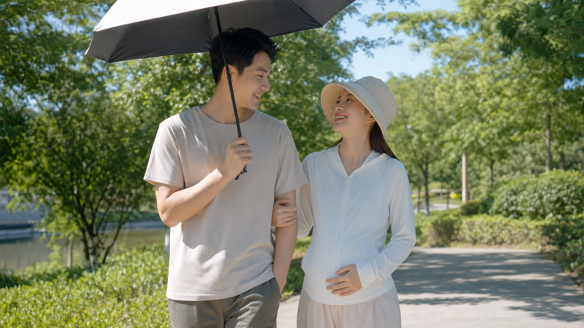 A loving Chinese couple walking outdoors in summer. The husband is holding a sun umbrella to protect his pregnant wife. The wife is wearing a stylish UPF sun protection jacket and a bucket hat, smiling happily. Sunny day, bright natural light, green park background, warm and affectionate atmosphere, realistic lifestyle photography, high quality, 8k.