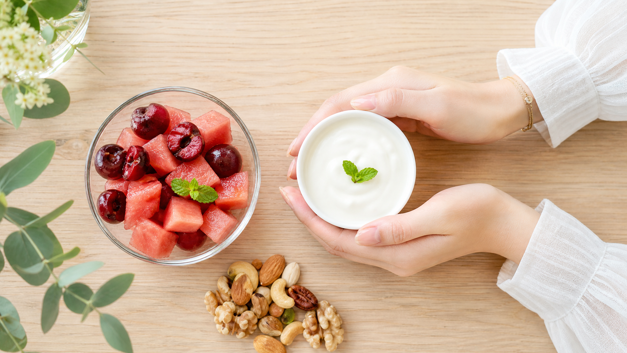 A top-down view of a healthy pregnancy snack on a light wooden table. A small glass bowl of diced watermelon and cherries (about the size of a fist), placed next to a cup of plain yogurt and a small handful of mixed nuts. A pair of elegant Chinese female hands is gently holding the yogurt cup. Bright and fresh color palette, food photography style, natural lighting, appetizing, highly detailed, 8k. --ar 3:4