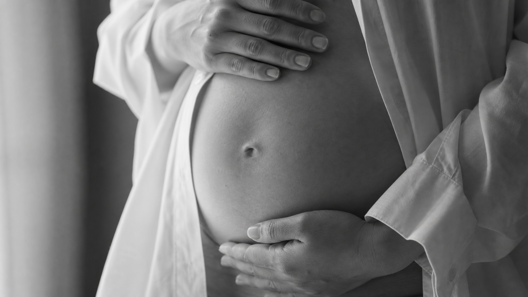 Close up of a Chinese pregnant woman's hands gently holding her bare baby bump. She is wearing an unbuttoned white shirt. Soft window light illuminating the contour of the belly. Black and white photography, high contrast, emotional, fine art photography, minimalist, highly detailed.