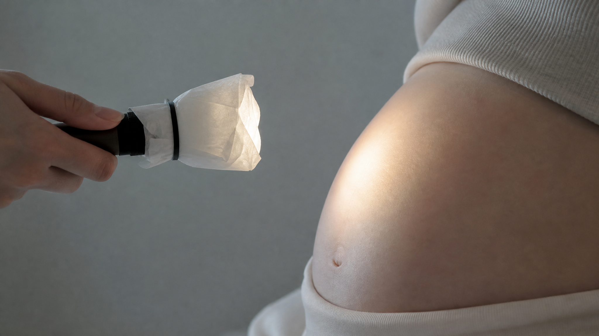 Close up of a Chinese pregnant woman's belly. A hand is holding a small flashlight covered with a layer of thin white tissue paper to soften the light. The flashlight is held at a safe distance of 15-20 cm away from the side of the belly. Soft grey and moon-white color palette, gentle gradient light beam, educational and safe atmosphere, photorealistic, macro photography, 8k.