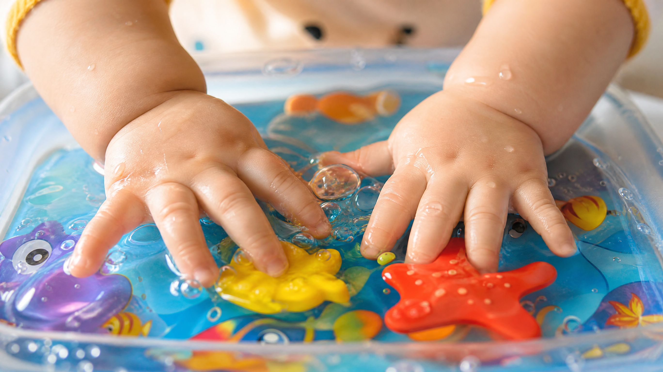 Close-up shot of a chubby Chinese baby's hands pressing on a transparent water play mat, colorful plastic sea creatures floating inside. Soft natural sunlight hitting the water surface, creating gentle reflections. High detail, macro photography, warm and inviting atmosphere.