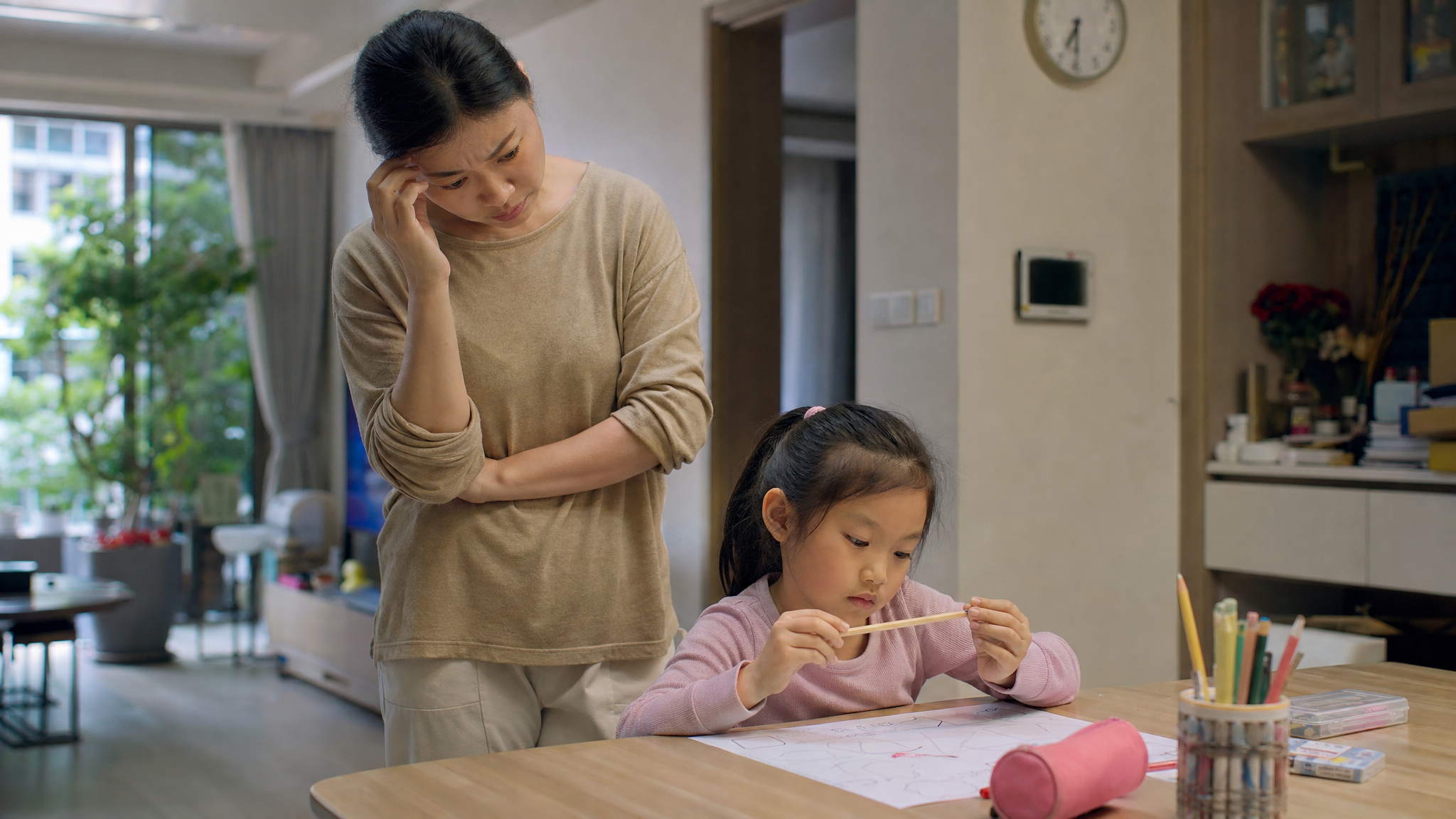 A high-quality, realistic photo of a frustrated Chinese mother standing next to her 5-year-old child. The child is sitting at a desk, looking distracted and playing with a pencil instead of drawing. The mother looks tired but loving. Soft indoor lighting, cozy modern Chinese apartment background. Shot on 35mm lens, cinematic lighting, 8k resolution. --ar 3:4