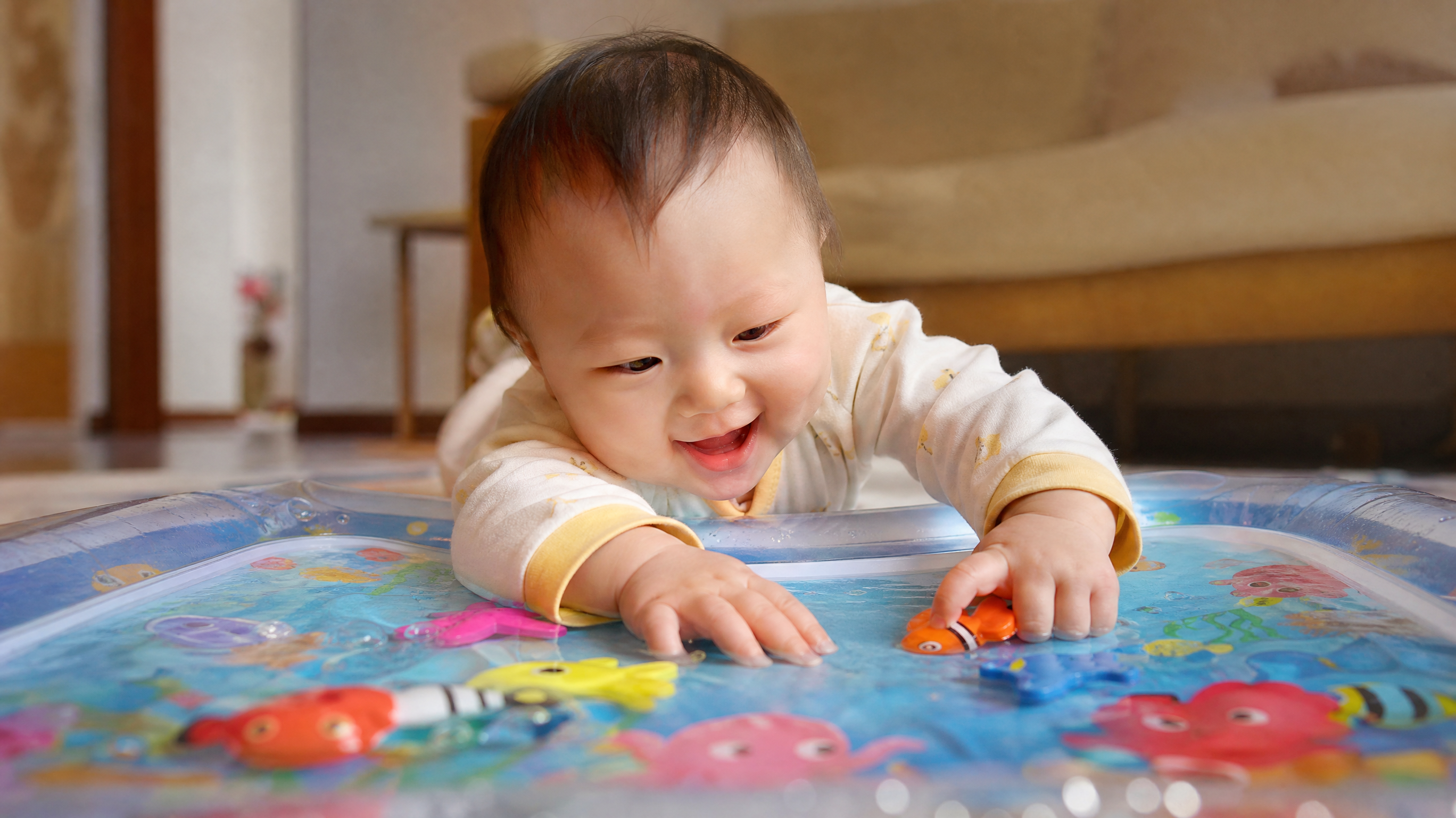 A cute Chinese baby lying on their tummy on a colorful water play mat, smiling happily, trying to catch floating toy fishes inside the mat. Bright and soft indoor lighting, cozy living room background, hyper-realistic, 8k resolution, photography, soft warm tones.