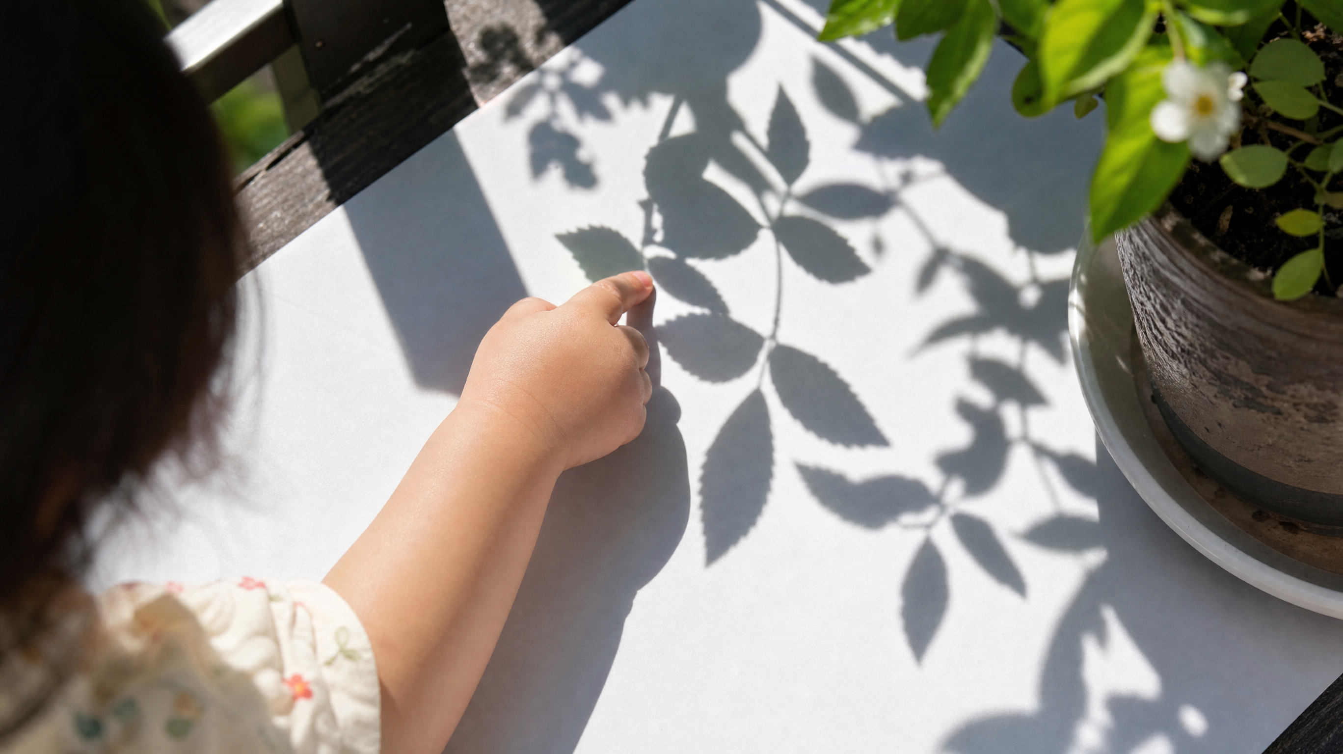 A top-down view of a 2-year-old Chinese toddler's little hand tracing the edge of beautiful leaf shadows cast on a white piece of paper on a sunlit balcony. High contrast between light and shadow, warm summer natural lighting, macro photography, highly detailed, aesthetic and peaceful vibe.