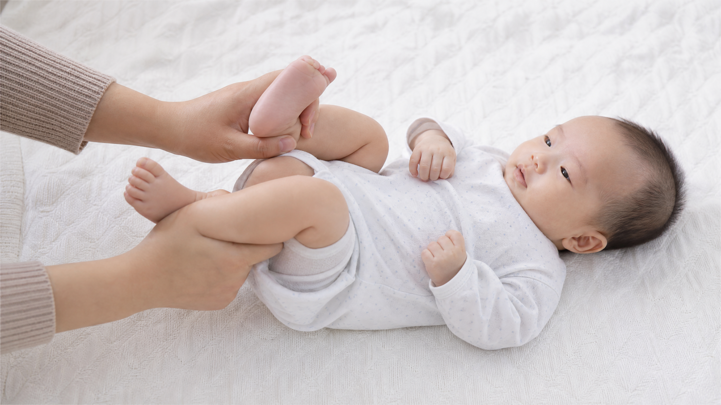 Close-up shot, top-down angle. A young Chinese mother's hands are gently holding the legs of a 2-month-old Chinese baby, doing the 
