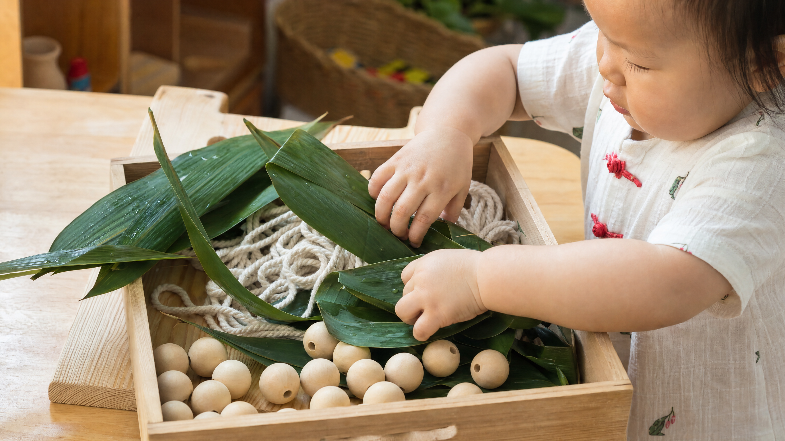 A high-quality, bright and warm photo of a sensory tray on a wooden table. Inside the tray, there are fresh green bamboo leaves (Zongzi leaves), thick cotton ropes, and large wooden beads. A cute 1-year-old Chinese toddler's chubby hands are reaching into the tray, playing with the leaves. Soft natural lighting from a window, cozy home environment, photorealistic, 8k resolution, shot on 35mm lens.