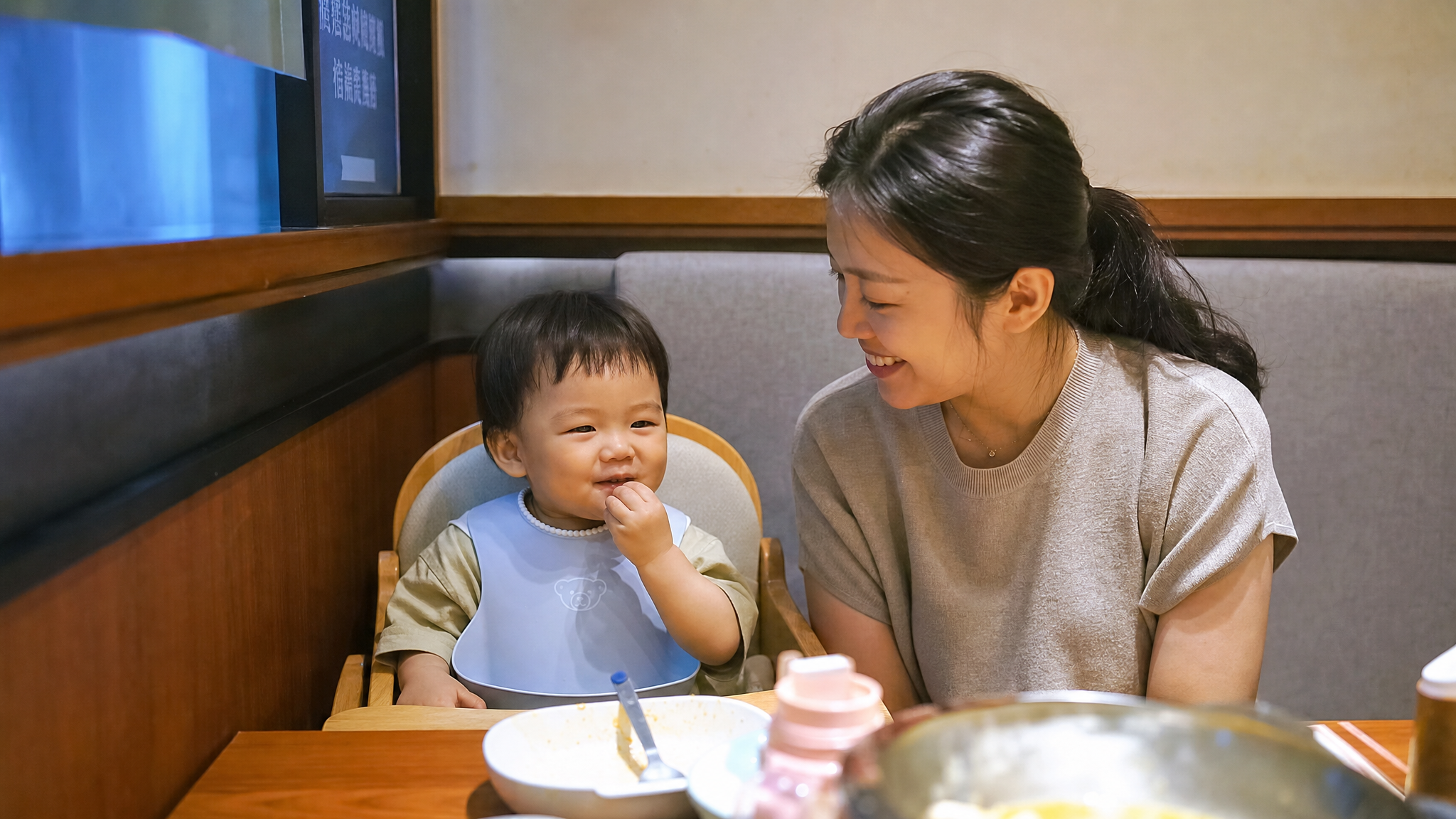 A warm and realistic photo of a young Chinese mother and her 1.5-year-old toddler sitting in a cozy corner booth of a restaurant. The baby is sitting quietly in a high chair next to the wall, away from the aisle. Soft indoor lighting, high resolution, photorealistic, shot on 35mm lens, joyful atmosphere.