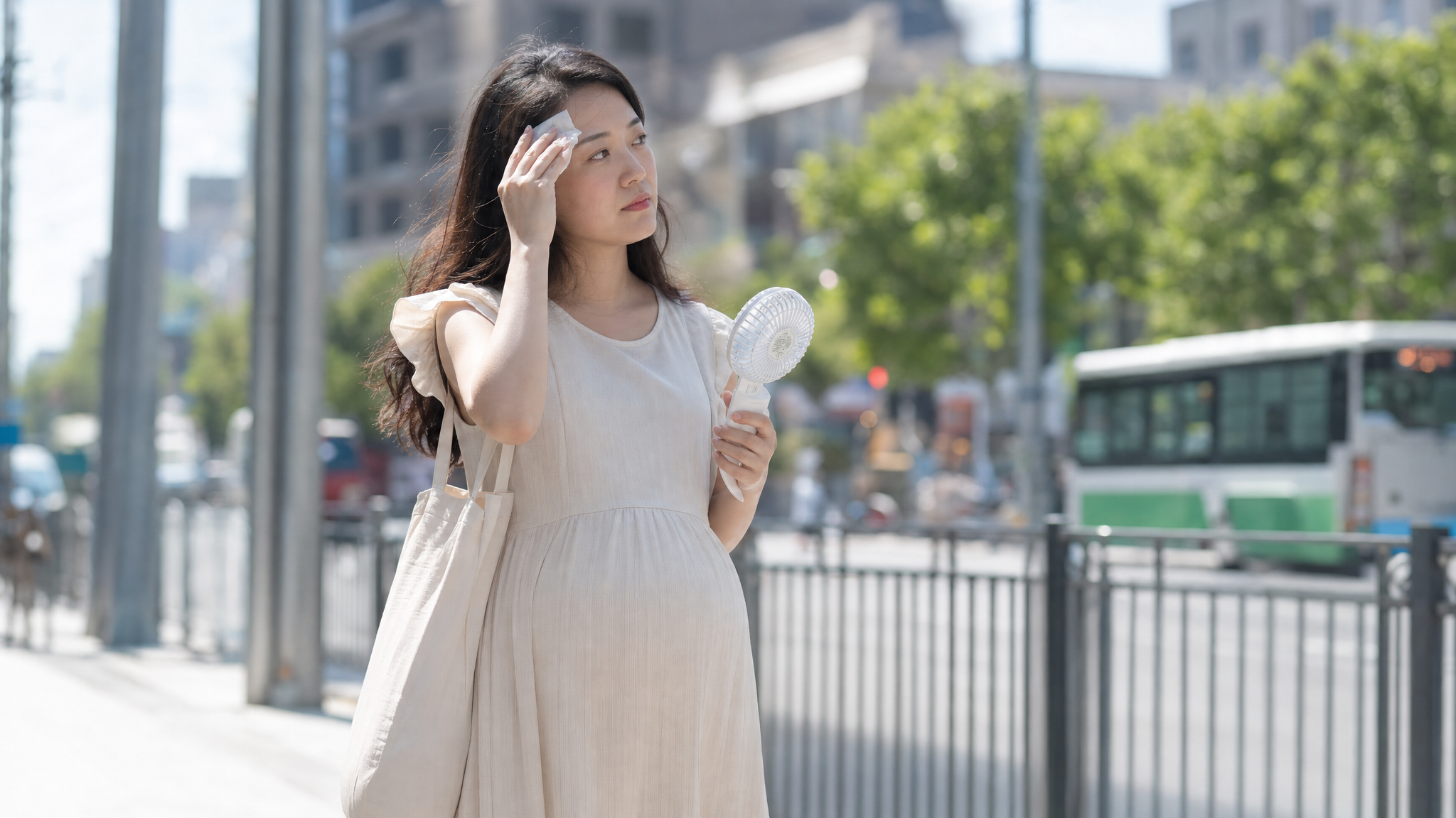 A realistic photography of a young Chinese pregnant woman commuting in summer, wearing a light-colored maternity dress, holding a small portable fan, gently wiping sweat from her forehead with a tissue. Bright sunlight, urban street background, soft lighting, 8k resolution, highly detailed. --ar 3:4