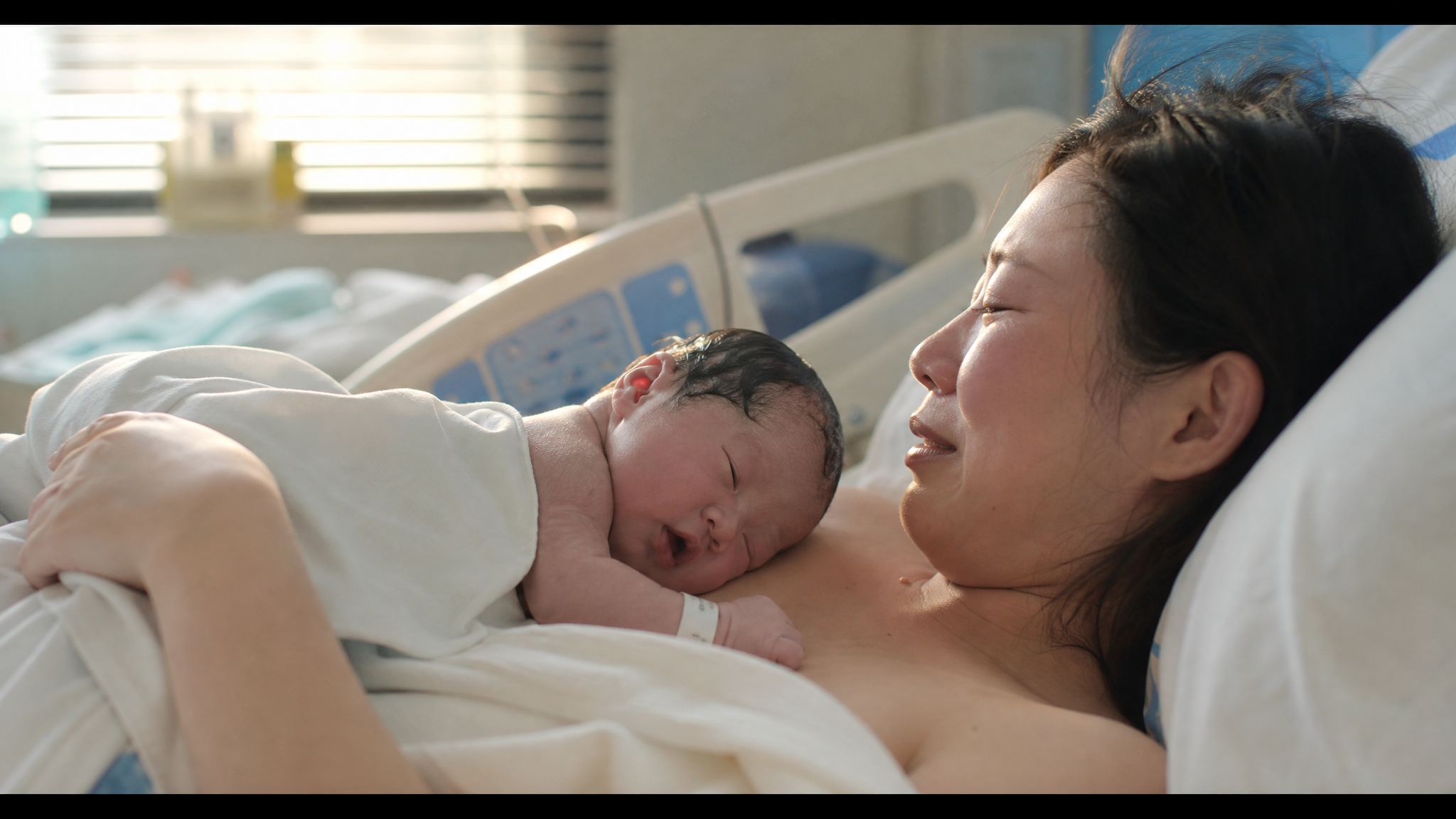 A heartwarming close-up shot of a Chinese newborn baby lying on the mother's bare chest in a softly lit hospital room. Morning sunlight gently filtering through window blinds. The mother is looking at the baby with tender, tearful eyes. Soft backlight, white hospital bedding with light blue accents. Warm, healing, cinematic lighting, photorealistic, shot on 35mm lens, 8k resolution. --ar 3:4 --v 6.0
