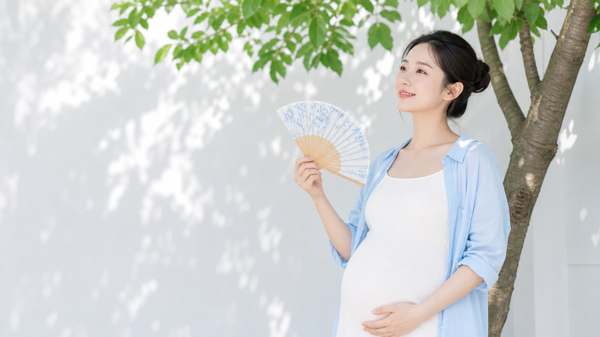 一位美丽的中国年轻孕妇,穿着清爽的蓝白配色夏季孕妇装,站在夏日阴凉的树荫下,手里拿着一把小巧的折叠扇