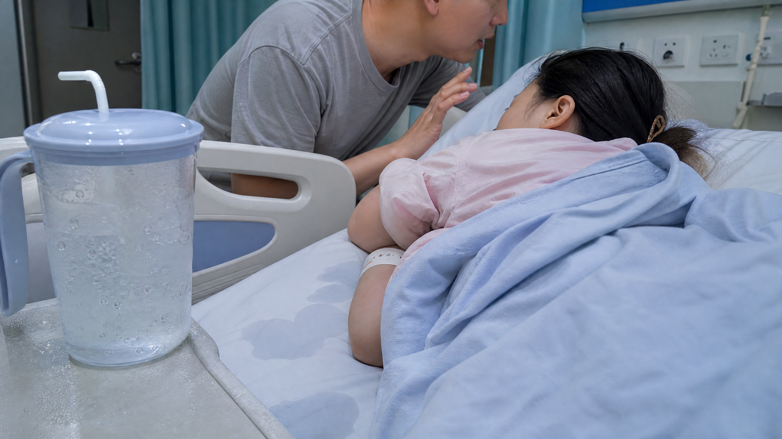 中国年轻产妇在医院产房床上侧躺休息，身上盖着薄毯，旁边中国年轻丈夫微微俯身靠近，用低声沟通的姿态安抚