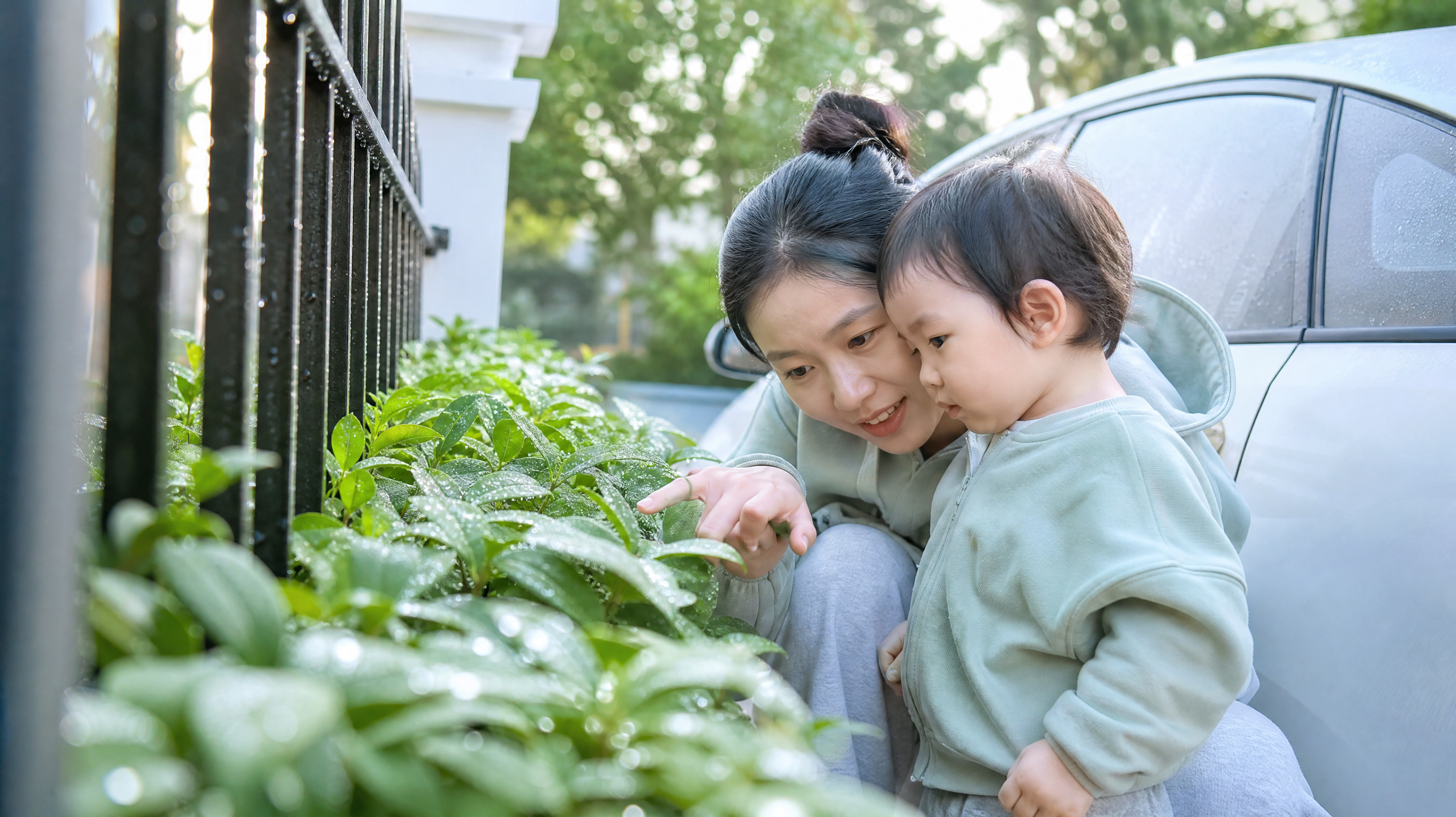 清晨小区楼下或窗边,柔和自然晨光;中国年轻妈妈半蹲陪中国1-3岁幼儿一起观察叶片和栏杆上的露珠,妈妈