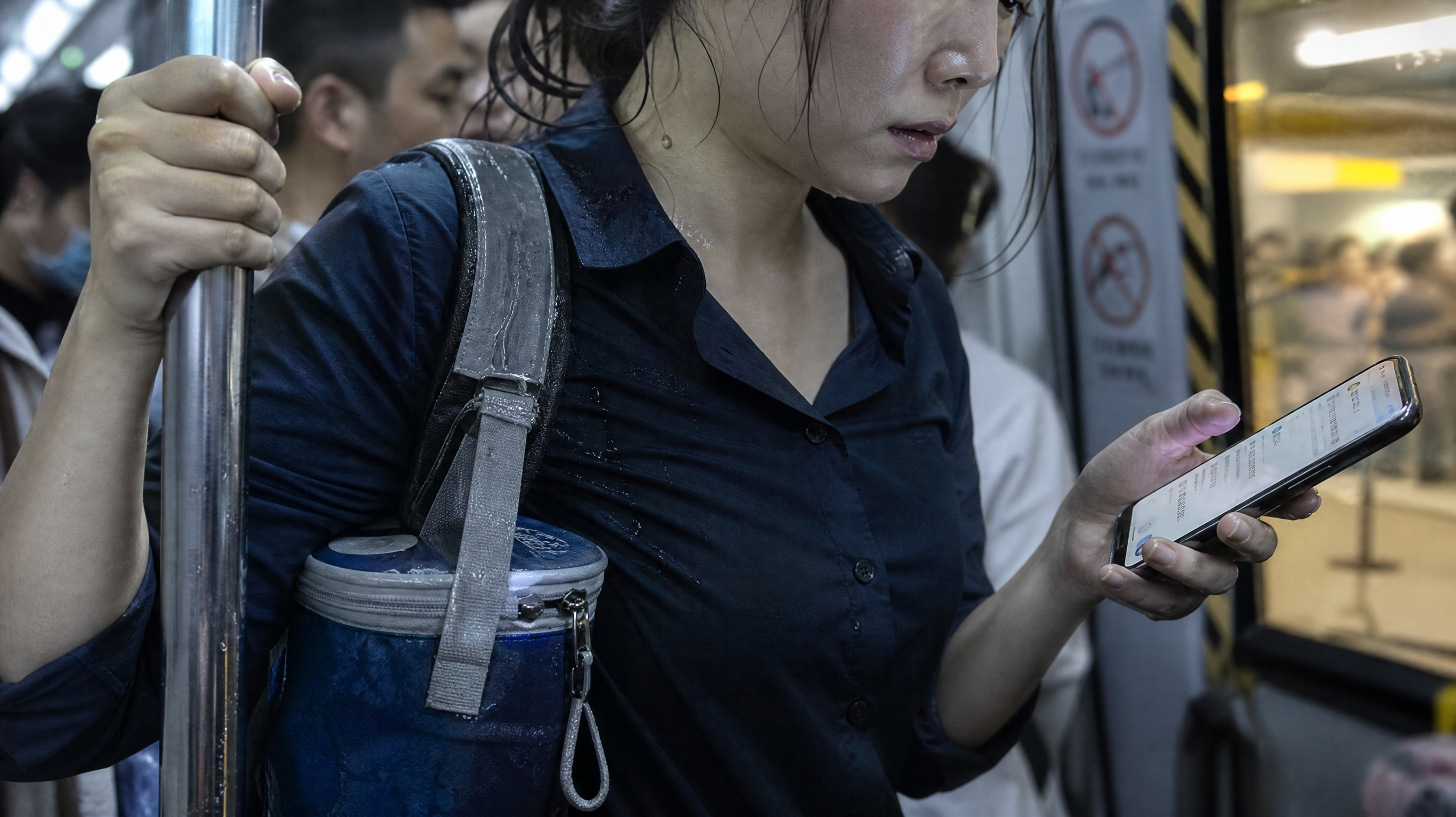 中国年轻职场妈妈在早高峰地铁车厢内的近景特写，肩上背包侧边挂着冰包，冰包冷凝水沿肩带滴落；妈妈一只手